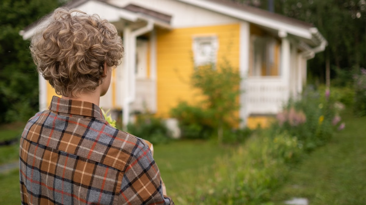 Teen with curly hair in plaid shirt stands facing a charming yellow house amidst lush greenery.